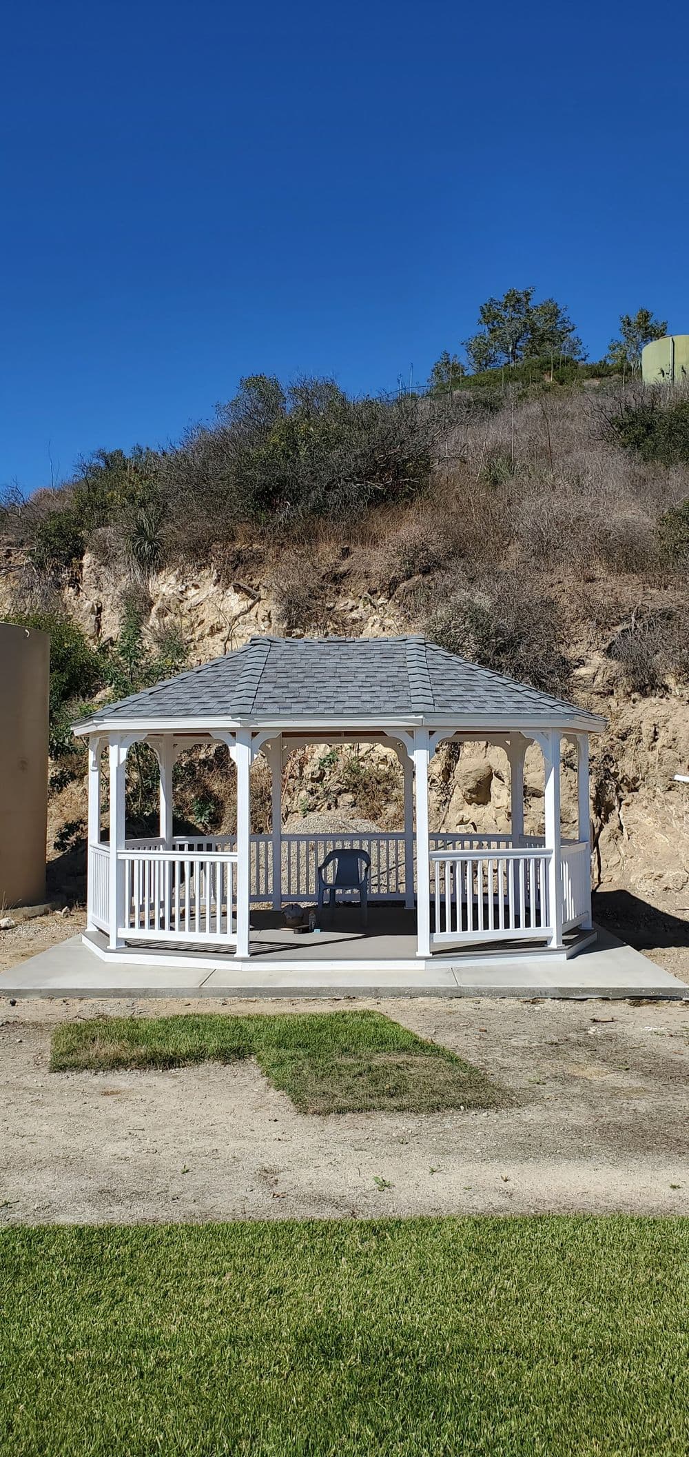 White gazebo on a grassy area with a hillside background under clear blue sky.