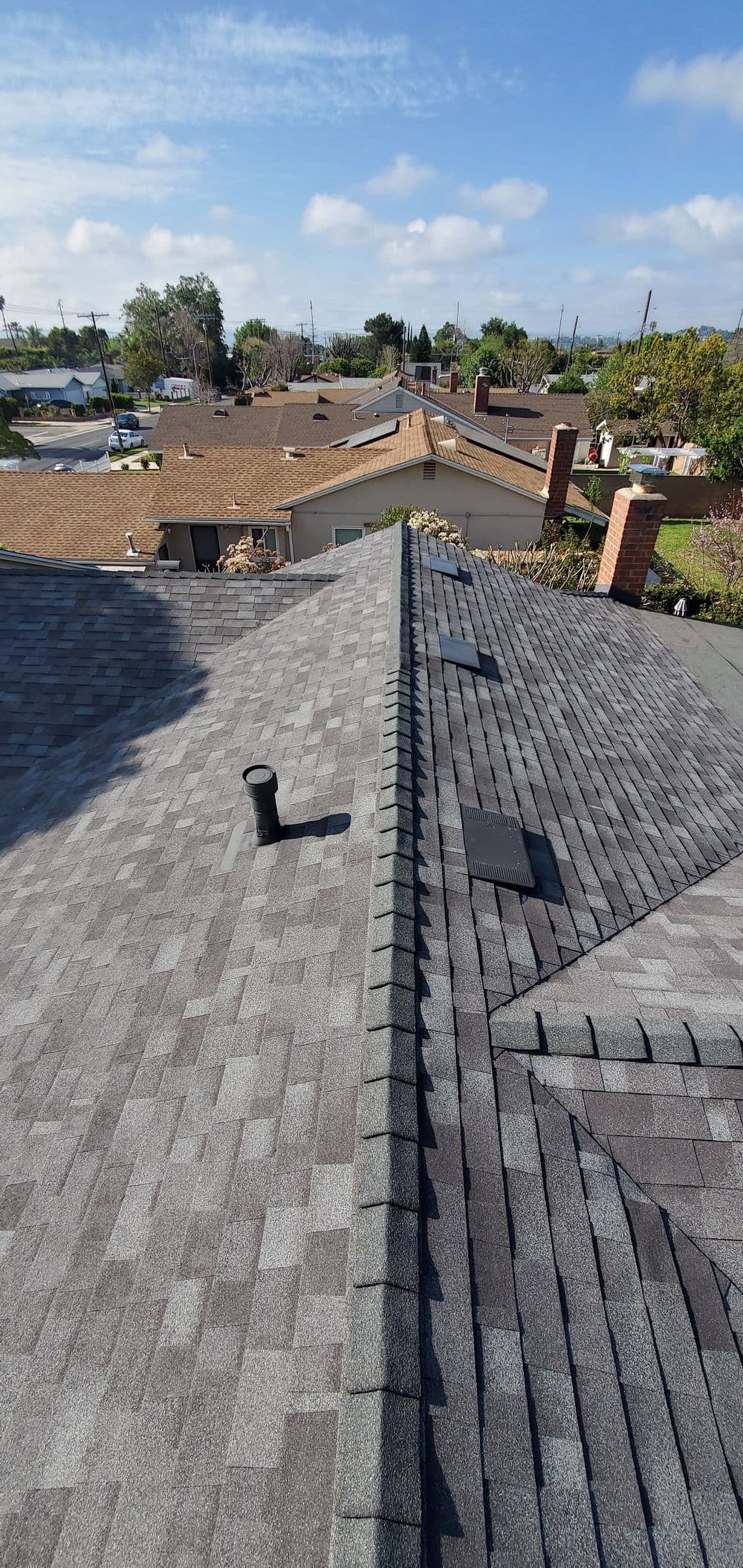 Aerial view of asphalt shingle roofs with chimney and vent under a clear blue sky.