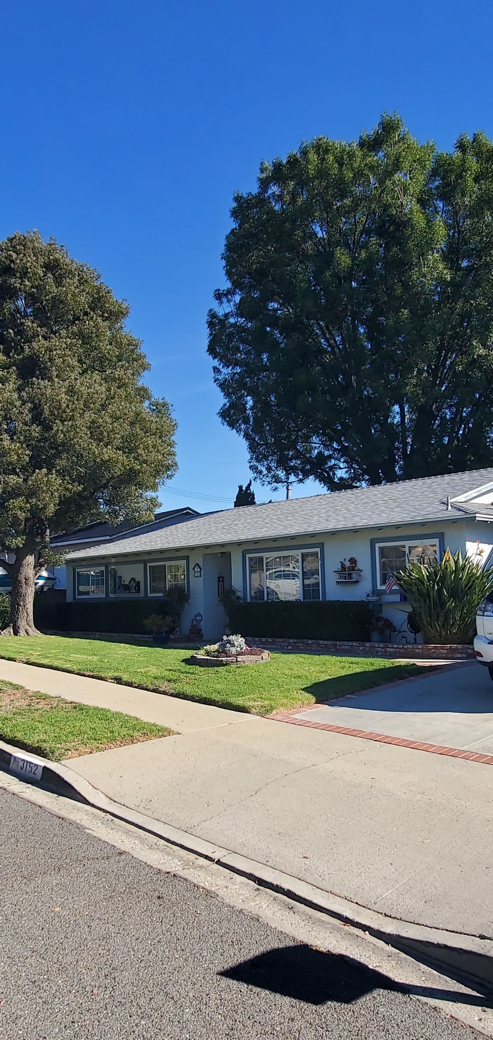 Single-story suburban home with a well-kept lawn and large trees under a clear blue sky.