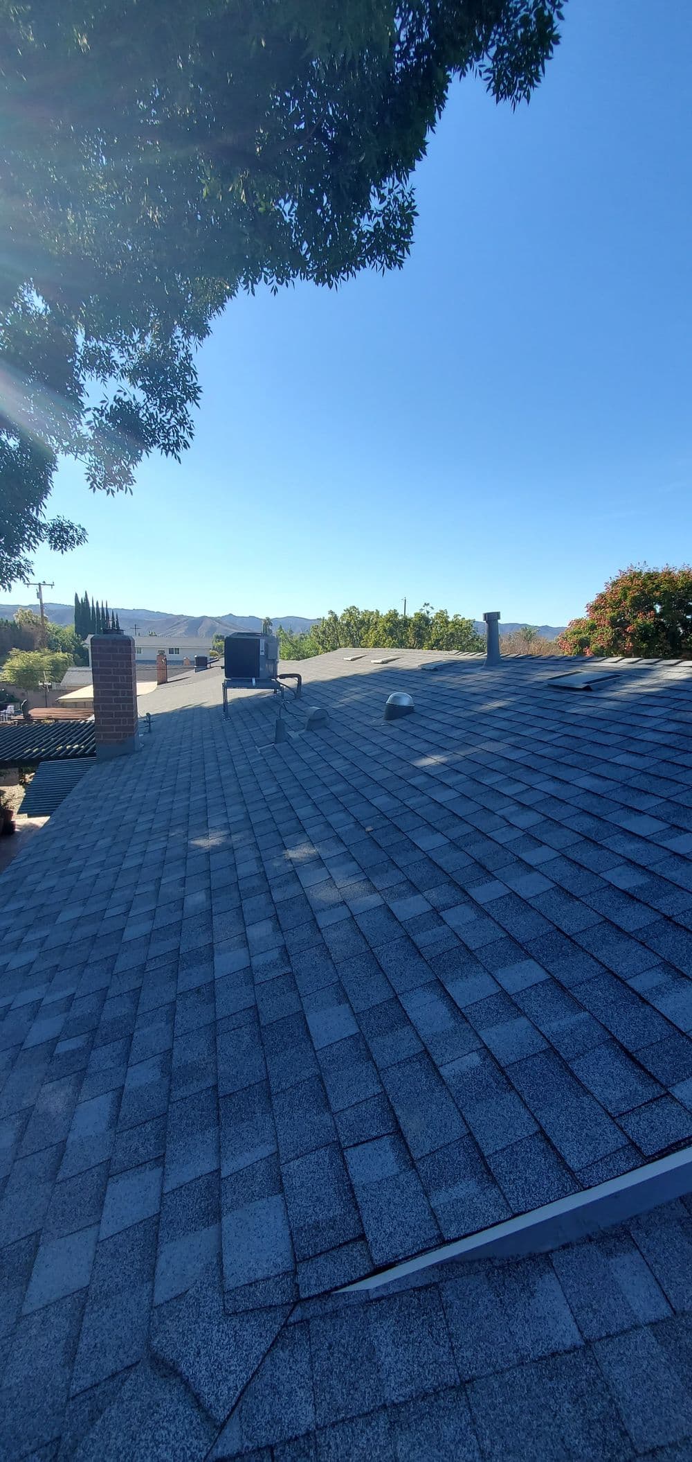 View of a residential roof with shingles, trees, and mountains in the background under clear blue sky.