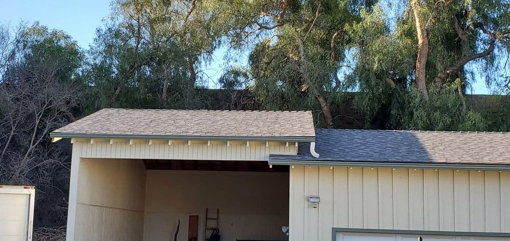 White garage with a grey roof beneath a canopy of trees. Clear blue sky in the background.