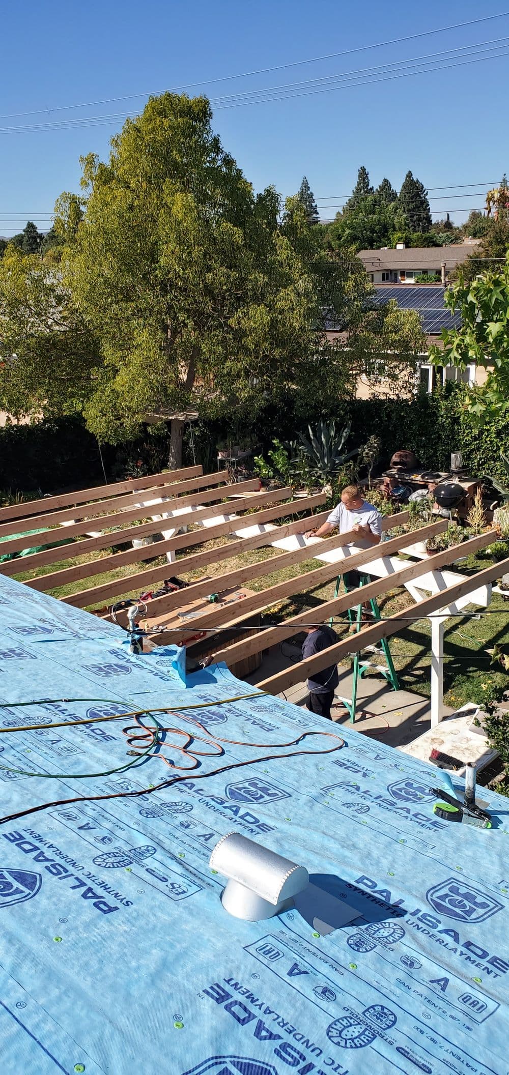 Construction workers installing a roof over a residential garden, with decking materials visible.