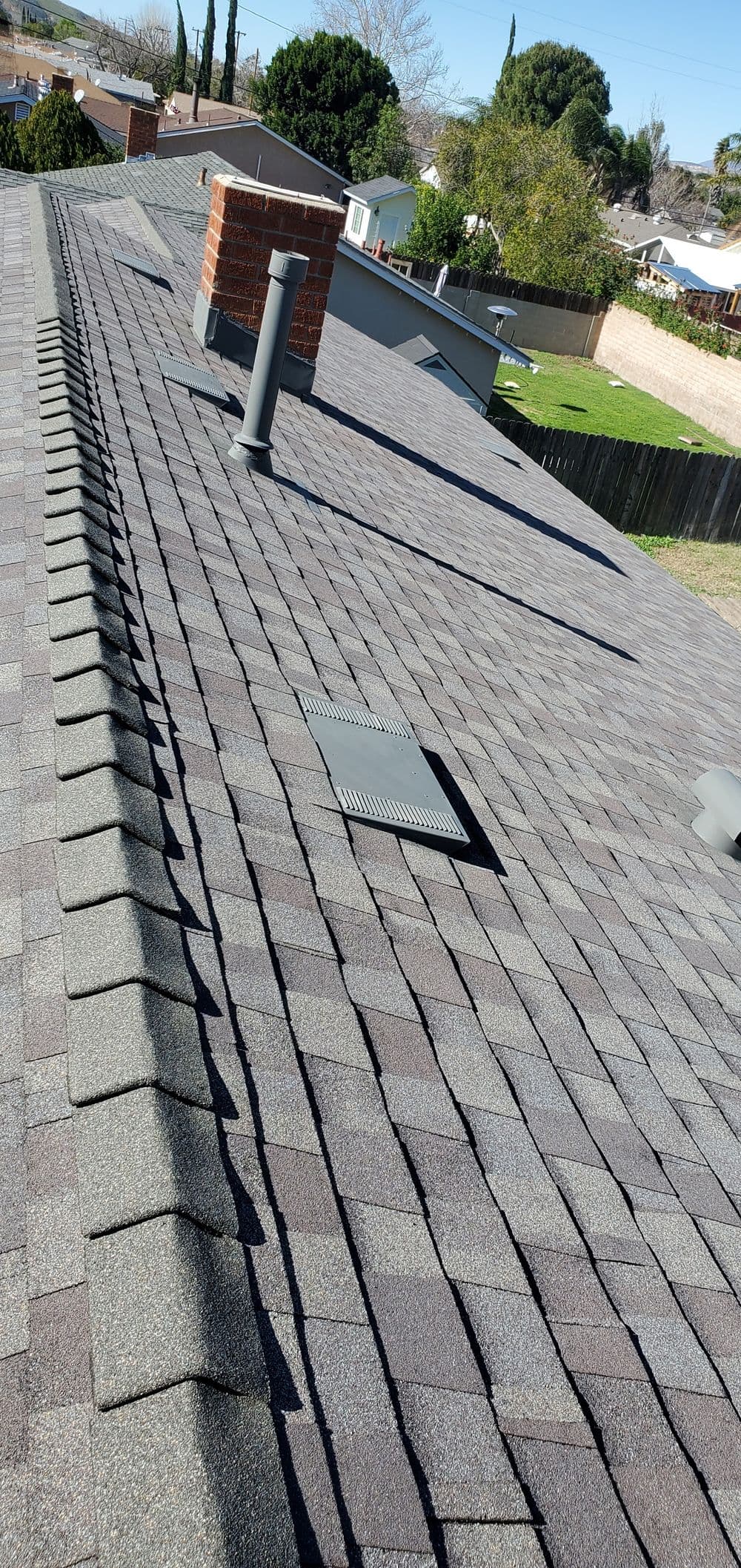 Aerial view of a gray shingle roof with vents and a chimney on a sunny day.